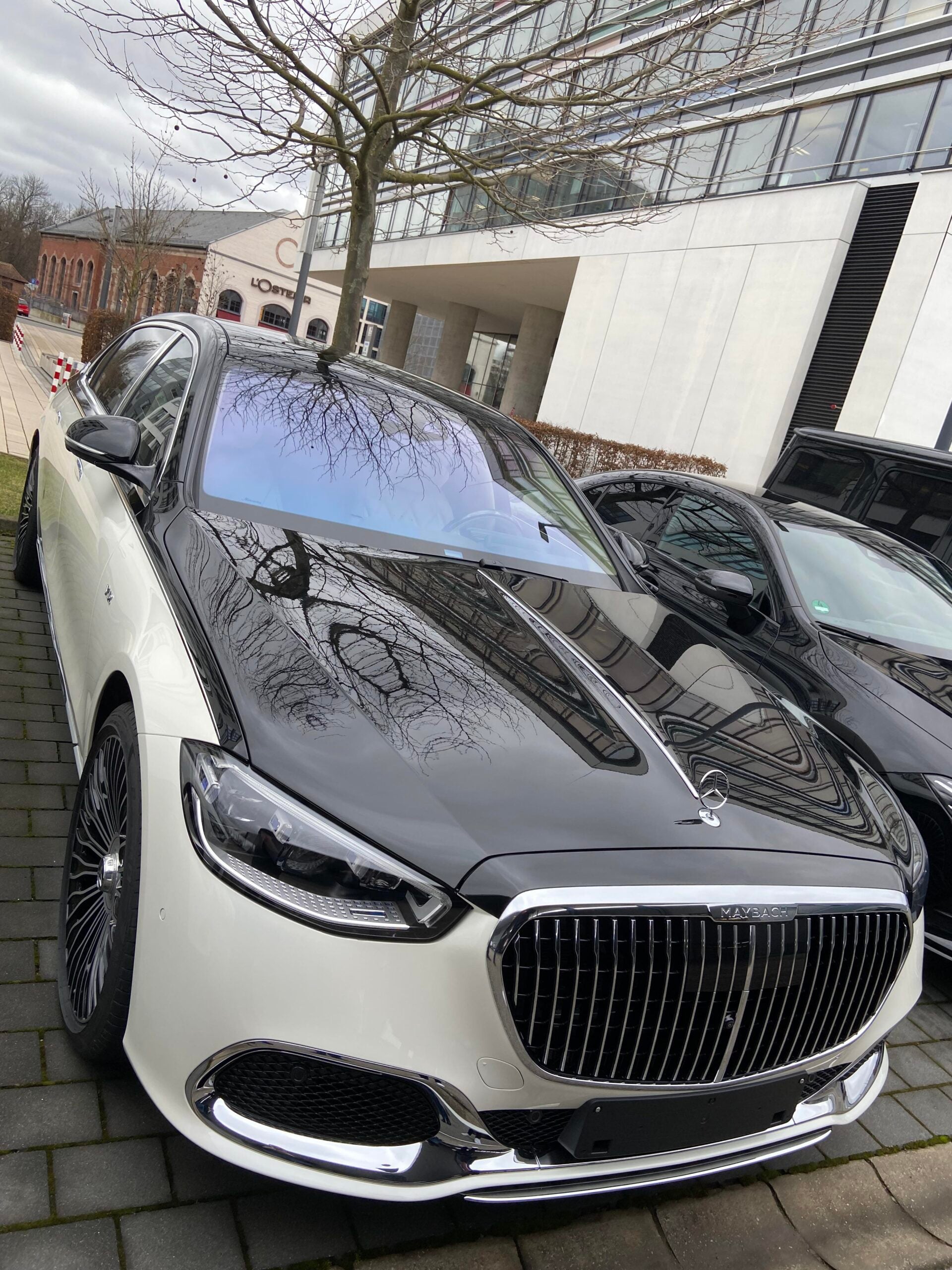 A black and white luxury sedan, likely a Mercedes-Maybach, is parked on a street in front of modern buildings with trees and another car nearby. The car features a large chrome grille and stylish rims.