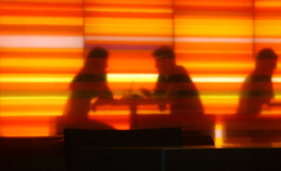 Silhouetted figures sit at tables in front of a glowing orange and yellow striped background, creating a warm, atmospheric scene. The image is slightly blurred, emphasizing the mood rather than the details.