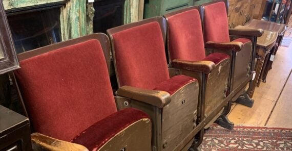 Four vintage theater seats with red velvet cushions and worn wooden armrests are lined up side by side on a wooden floor in what appears to be an antique shop.