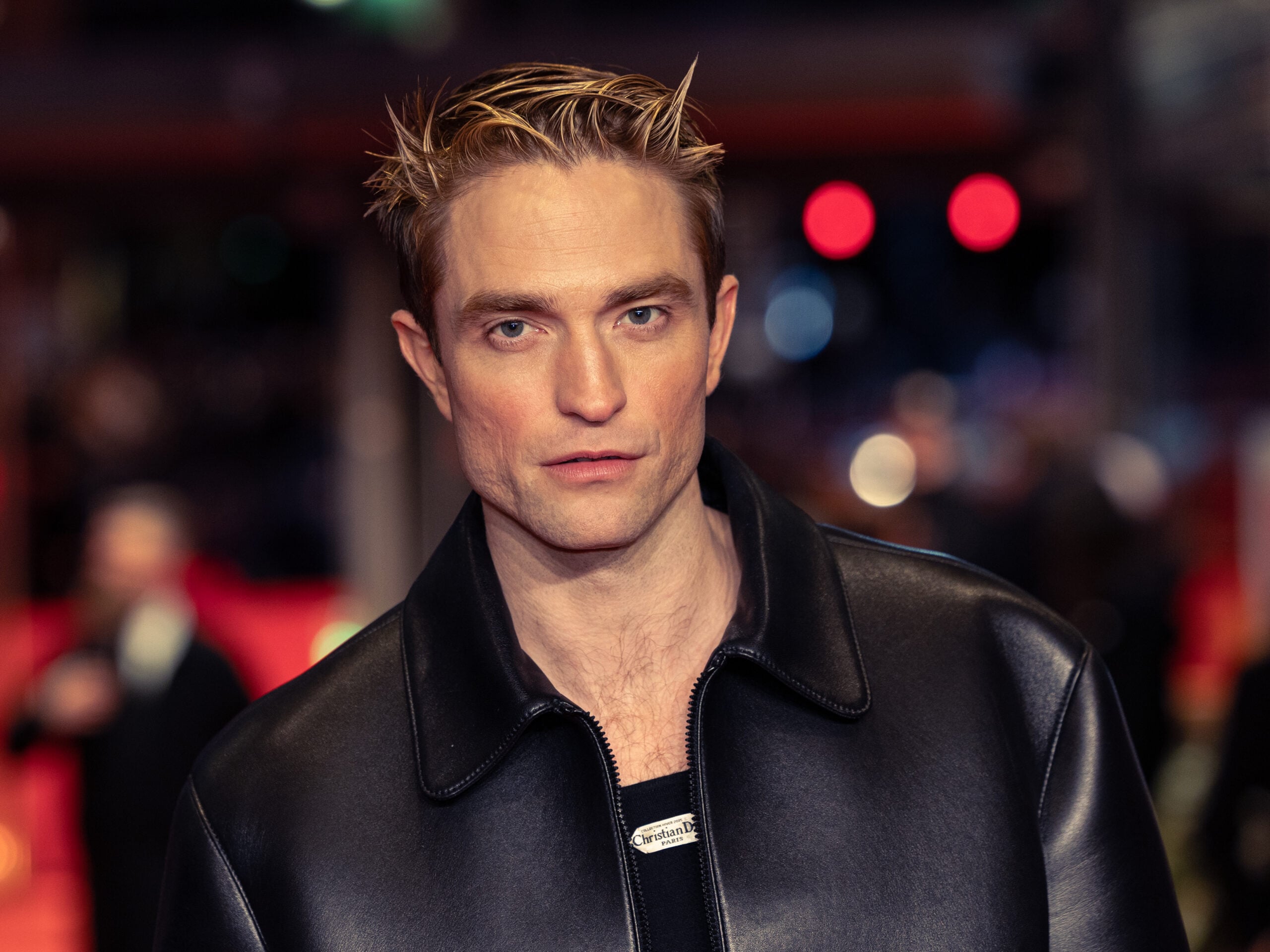 A man with short, spiked blond hair wearing a dark leather jacket stands in front of a blurred background with red and black tones and lights.