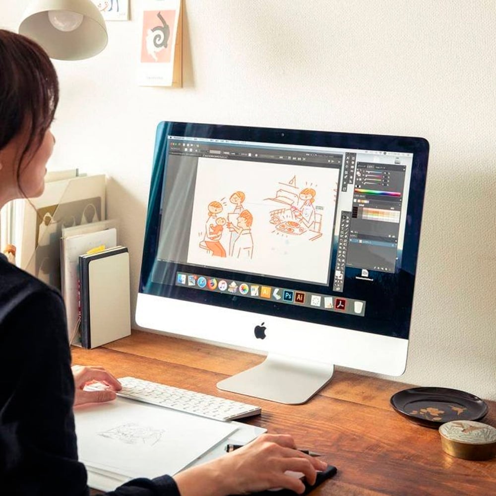 A person sits at a wooden desk using a large iMac computer to create a digital illustration of people at a table. Art supplies, books, and a keyboard are also on the desk.