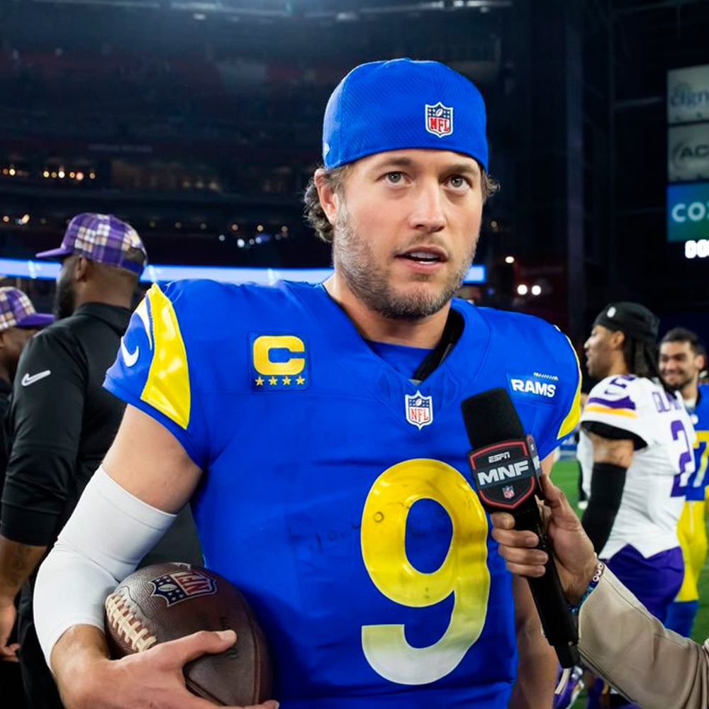 A Los Angeles Rams football player in uniform, wearing a blue cap backward, holds a football while being interviewed on the field after a game. Other players and reporters are visible in the background.