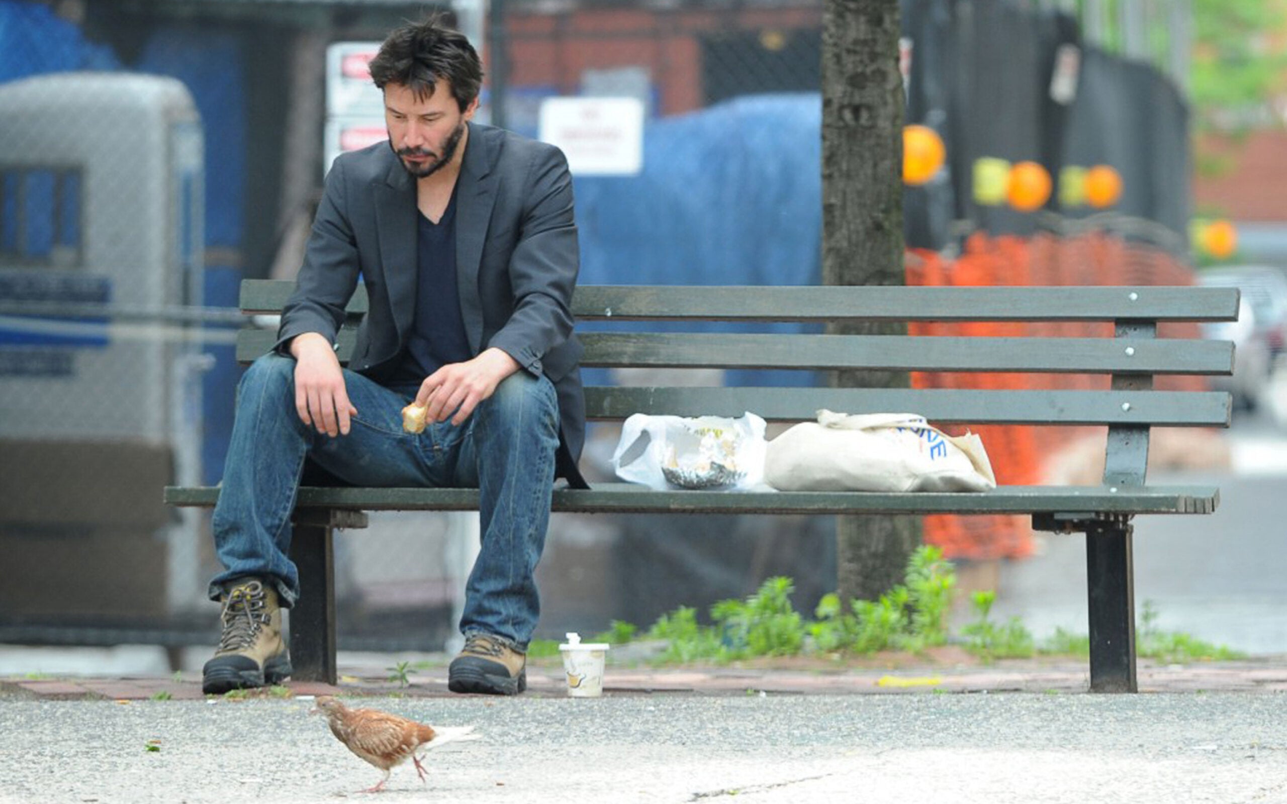 A man in a dark jacket and boots sits alone on a park bench, holding food. A paper bag and drink are beside him. He looks down while a small bird stands on the ground in front of him. The background is urban.
