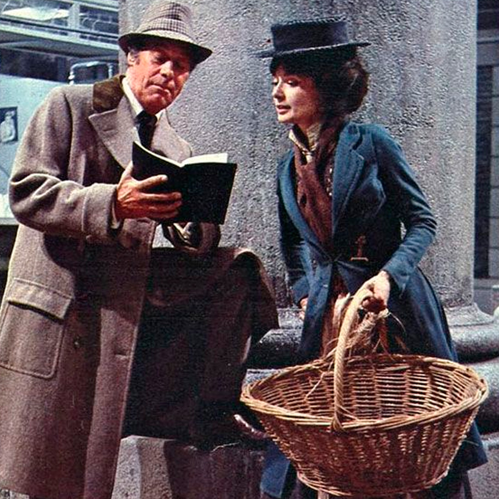 A man in a gray overcoat and hat reads from a book beside a woman in a blue dress and hat holding a large empty basket. They stand next to a stone column in what appears to be an old-fashioned setting.