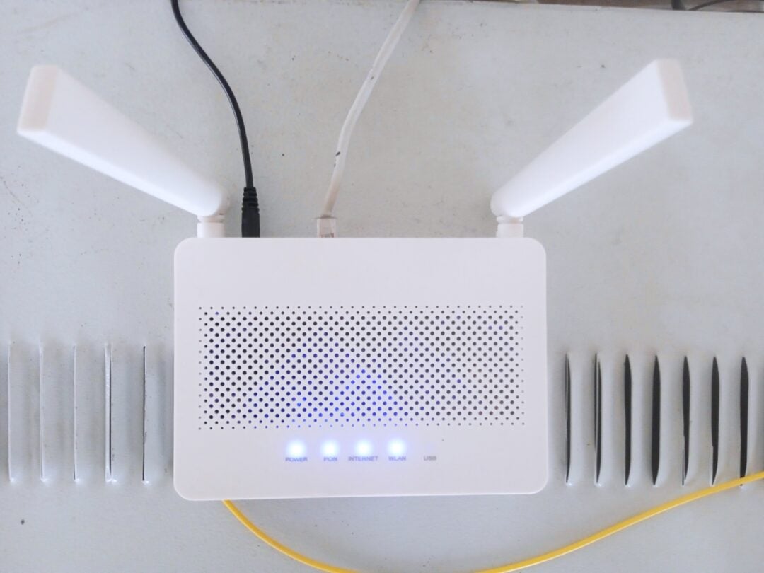 A white wireless router with two antennas, four blue indicator lights, and connected cables sits on a light-colored surface with rows of gray objects on each side.