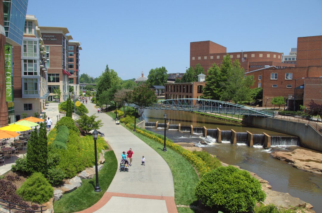 A sunny day at a riverside park with people walking on a paved path, outdoor dining tables with umbrellas, modern buildings, a small bridge, and a waterfall in the river. Trees and green lawns line the walkway.
