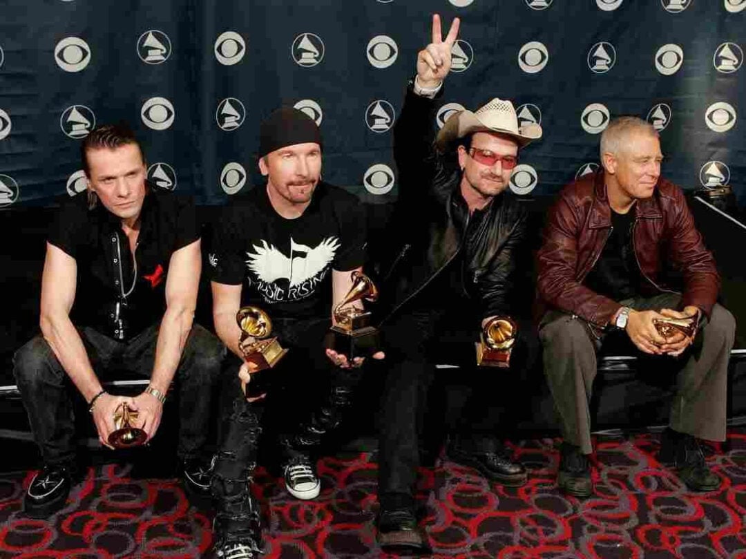 Four men sit on a black couch holding Grammy Awards, posing in front of a Grammy-branded backdrop. One man wears a hat and sunglasses, making a peace sign. All appear happy and relaxed.