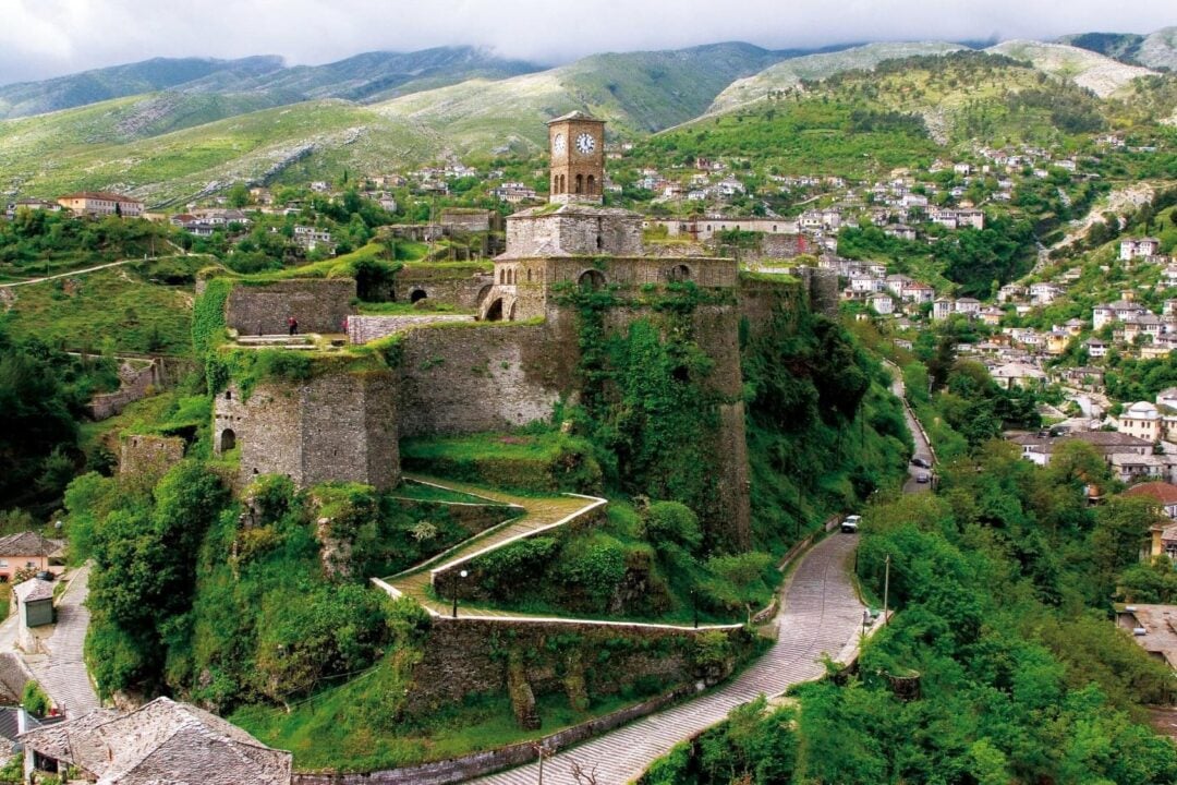 A historic stone castle with a clock tower sits atop a lush green hill, surrounded by winding roads and a village with scattered houses, with mountains visible in the background under a partly cloudy sky.