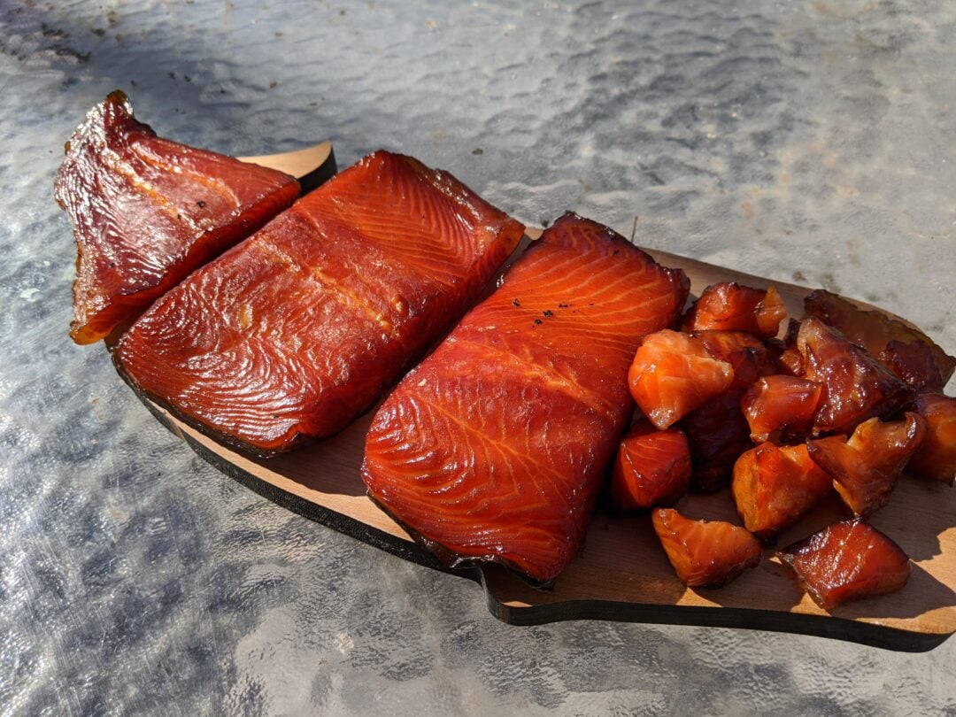 Slices and chunks of smoked salmon are arranged on a wooden board, with a shiny, reddish-brown surface, set on a textured glass table in natural sunlight.