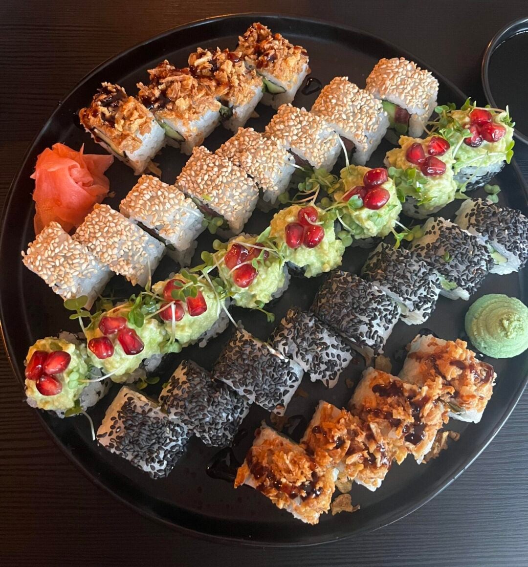 A round black plate with assorted sushi rolls, topped with sesame seeds, pomegranate seeds, crispy onions, and microgreens. Sides of pickled ginger and wasabi are on the plate, set on a dark table.