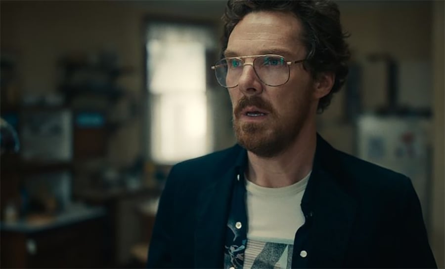 A man with curly brown hair, a beard, and glasses stands indoors, looking surprised or concerned. He is wearing a dark jacket over a printed t-shirt, and the background appears to be a dimly lit kitchen.