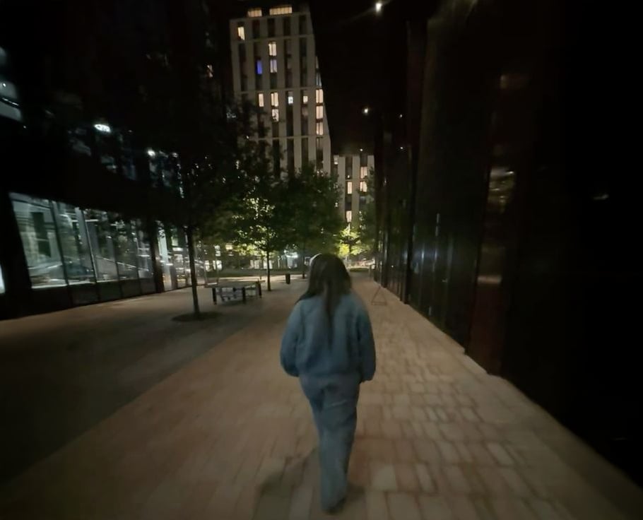 A person wearing a blue outfit walks alone on a dimly lit city sidewalk at night, surrounded by tall buildings with illuminated windows and a few trees along the path.