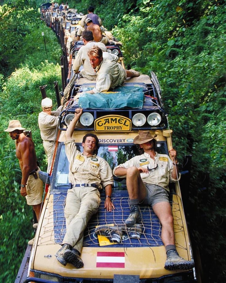 A group of people in safari-style clothing sit and lie on top of off-road vehicles marked "Camel Trophy," driving through dense green jungle foliage in a rugged adventure setting.