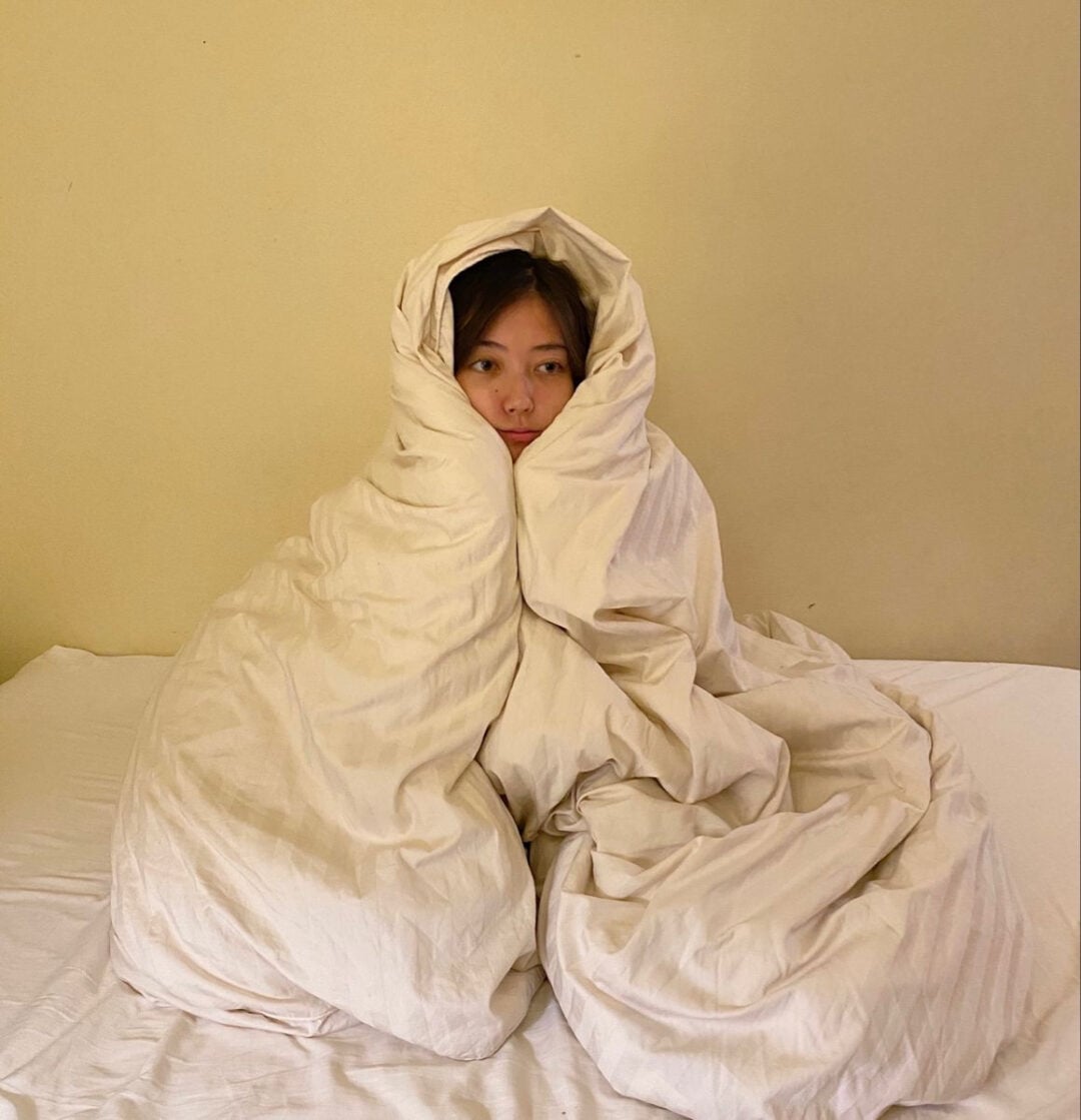 A person is sitting on a bed wrapped tightly in a beige blanket, with only their face visible, creating a cocoon-like appearance against a plain wall.