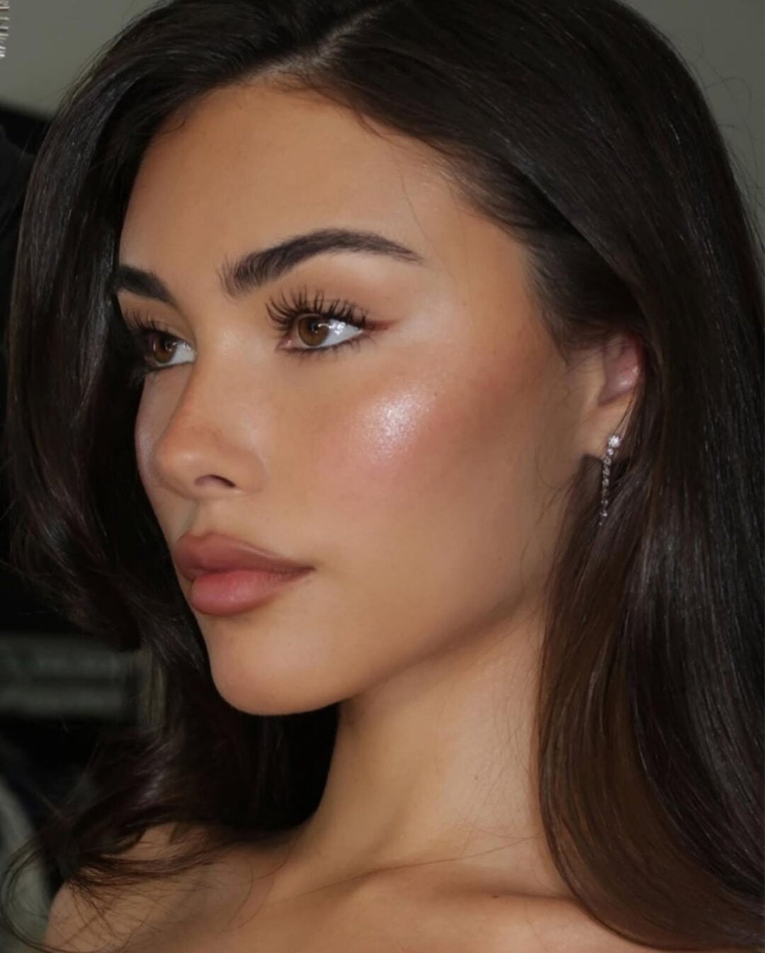 A young woman with long brown hair and glowing skin looks to the side. She has full lips, defined brows, long eyelashes, and wears a small hoop earring. The lighting highlights her smooth, dewy complexion.