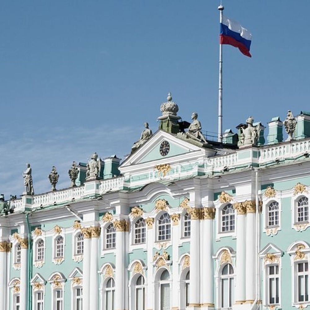 The facade of the Winter Palace in St. Petersburg, Russia, featuring ornate turquoise and white architecture with gold detailing, statues on the roof, and a Russian flag flying above.