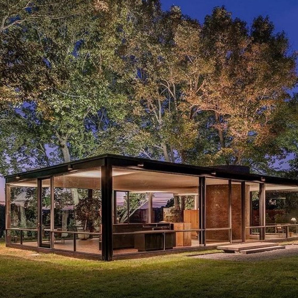 A modern glass house with black frames sits on a grassy lawn, surrounded by tall trees, illuminated warmly at dusk under a clear evening sky.