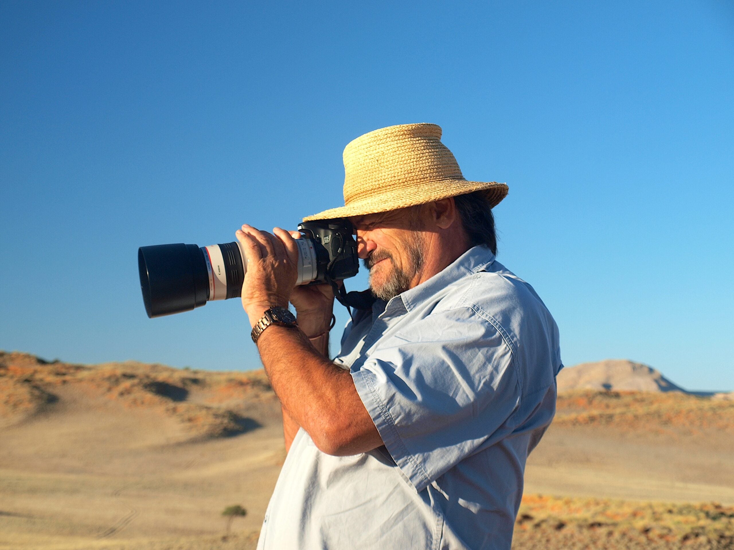 A man wearing a straw hat and light blue shirt is taking a photo with a camera fitted with a large lens, standing outdoors in a sunny, desert-like landscape with hills in the background.