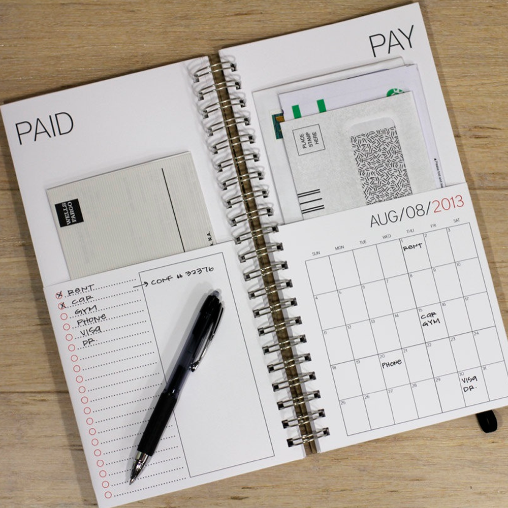An open planner on a wooden table shows a monthly calendar, bills, a to-do list with a pen, and sections labeled "PAID" and "PAY." The calendar is marked for August 2013 and some bills are tucked inside.
