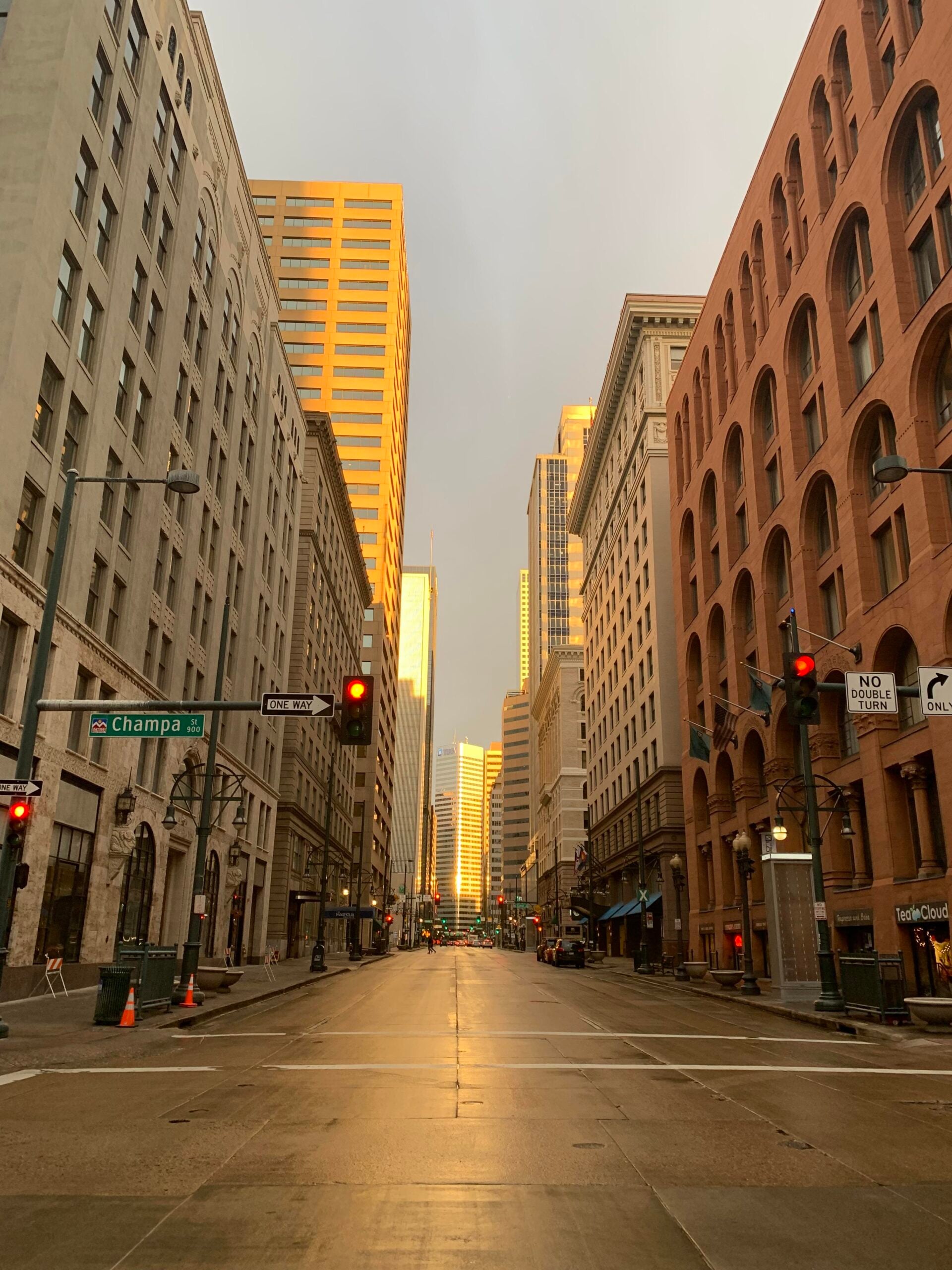 A city street at sunset, empty of cars and people, lined with tall buildings. The sunlight reflects warmly off the windows and wet pavement. Street signs for Champa St. and 17th St. are visible.