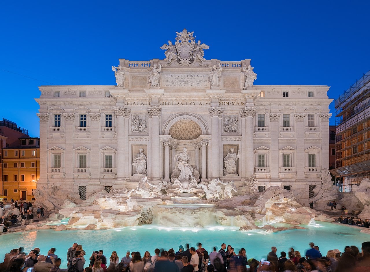 The Trevi Fountain in Rome is illuminated at dusk, with intricate statues and clear water in the foreground, and a crowd of people gathered along the edge of the fountain.