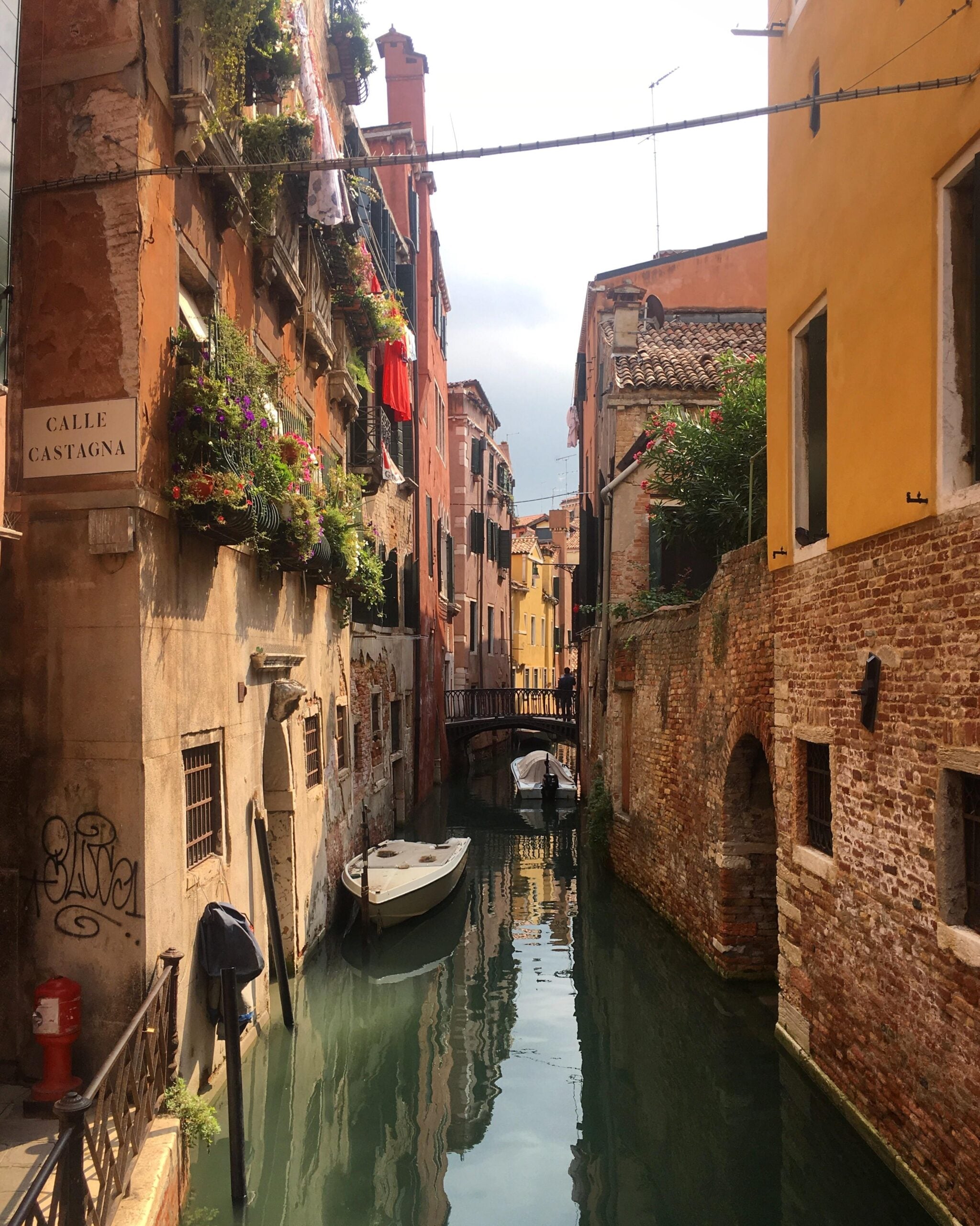 Narrow canal in Venice lined with colorful buildings, balconies with flowers, hanging laundry, and small boats moored along the water. A sign reads "Calle Castagna" and a small bridge crosses the canal in the distance.