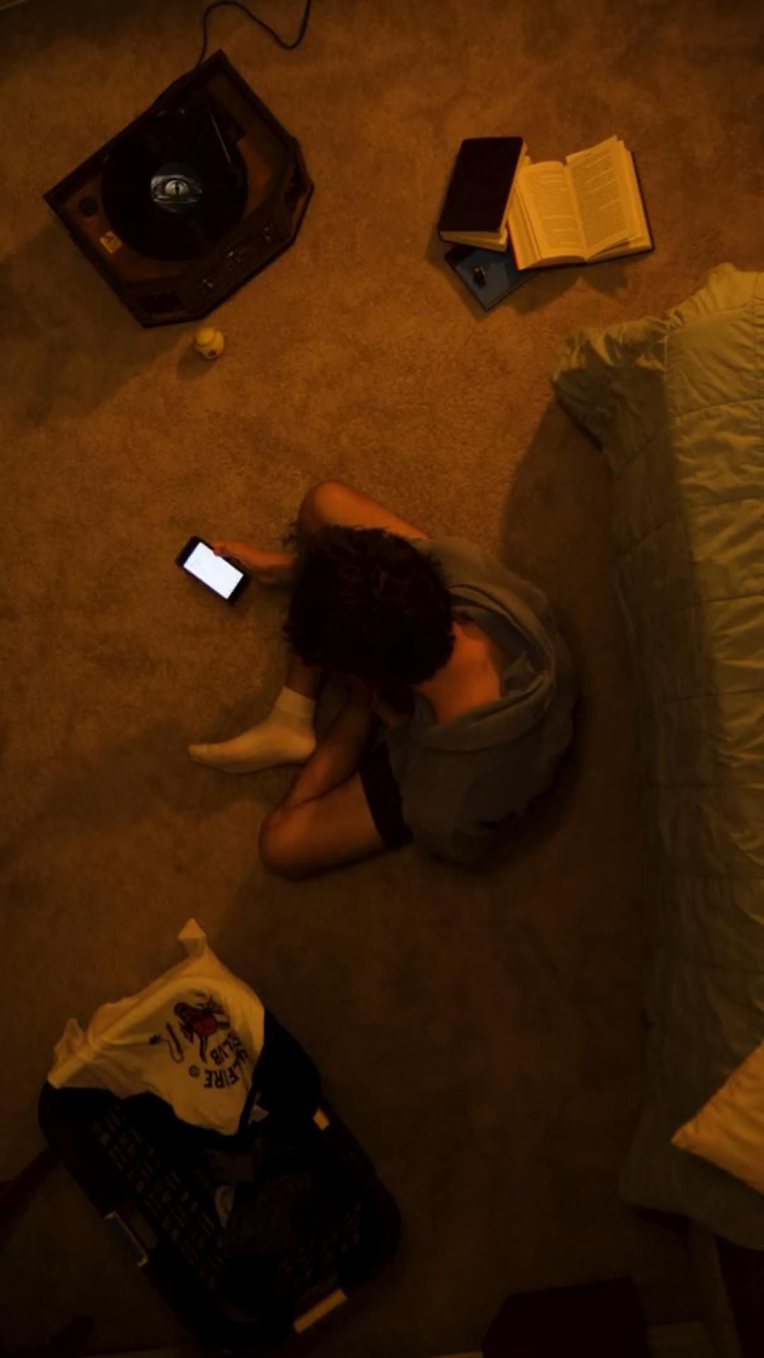 A person sits cross-legged on a carpeted floor, looking at a phone. Nearby are an open book, a record player, a laundry basket with clothes, and the edge of an unmade bed. The lighting is warm and dim.