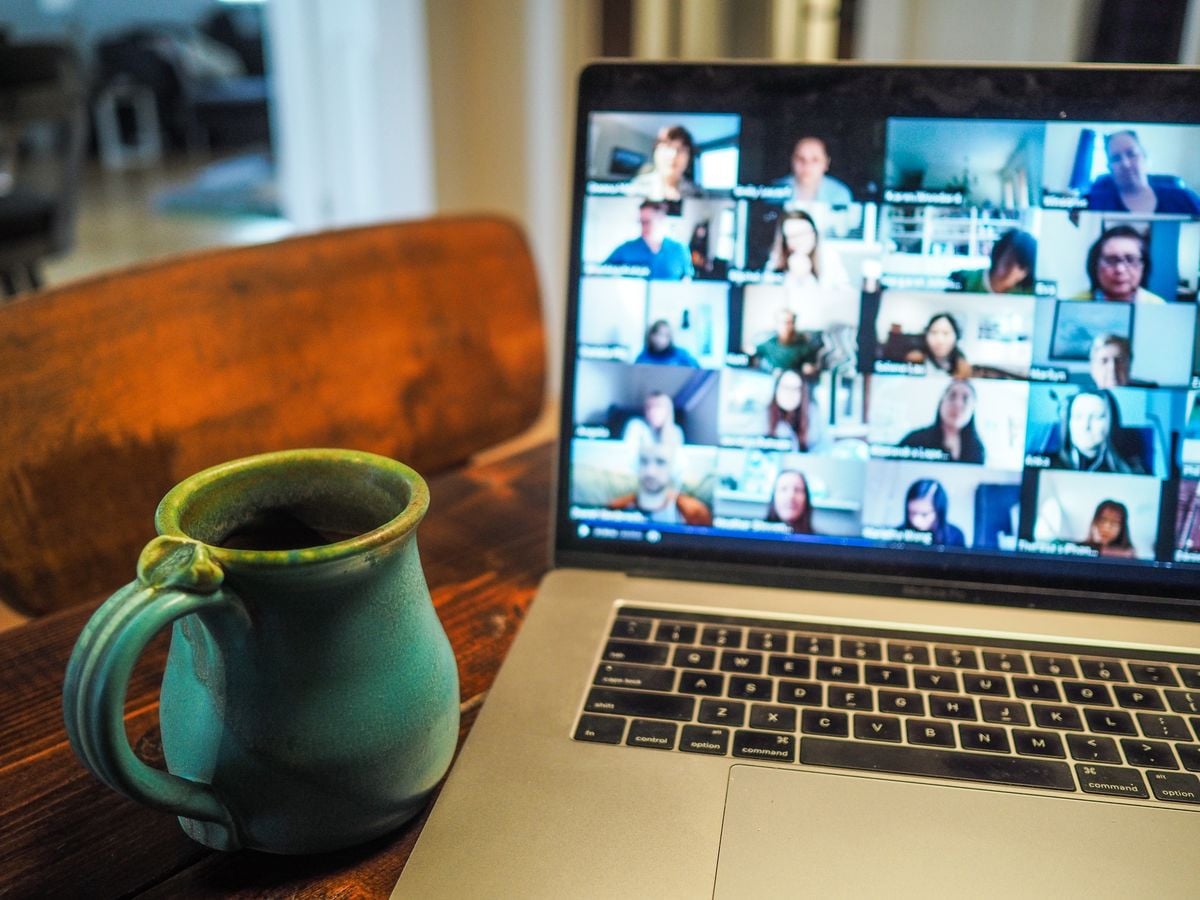A laptop on a wooden table displays a video conference with many participants, next to a green ceramic mug filled with a dark beverage. The background is softly blurred.