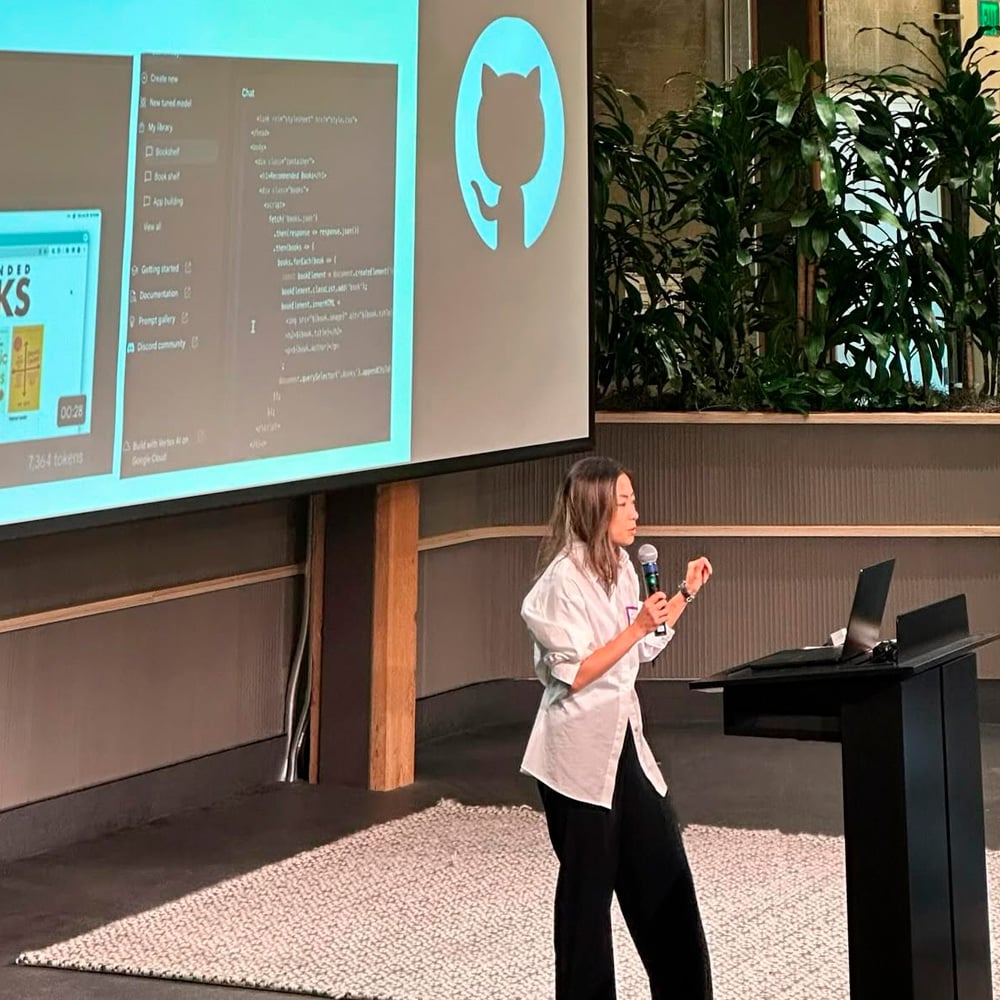 A woman is speaking at a podium with a laptop, gesturing with her hand. Behind her, a screen displays code and the GitHub logo. The room has plants and a carpeted area.