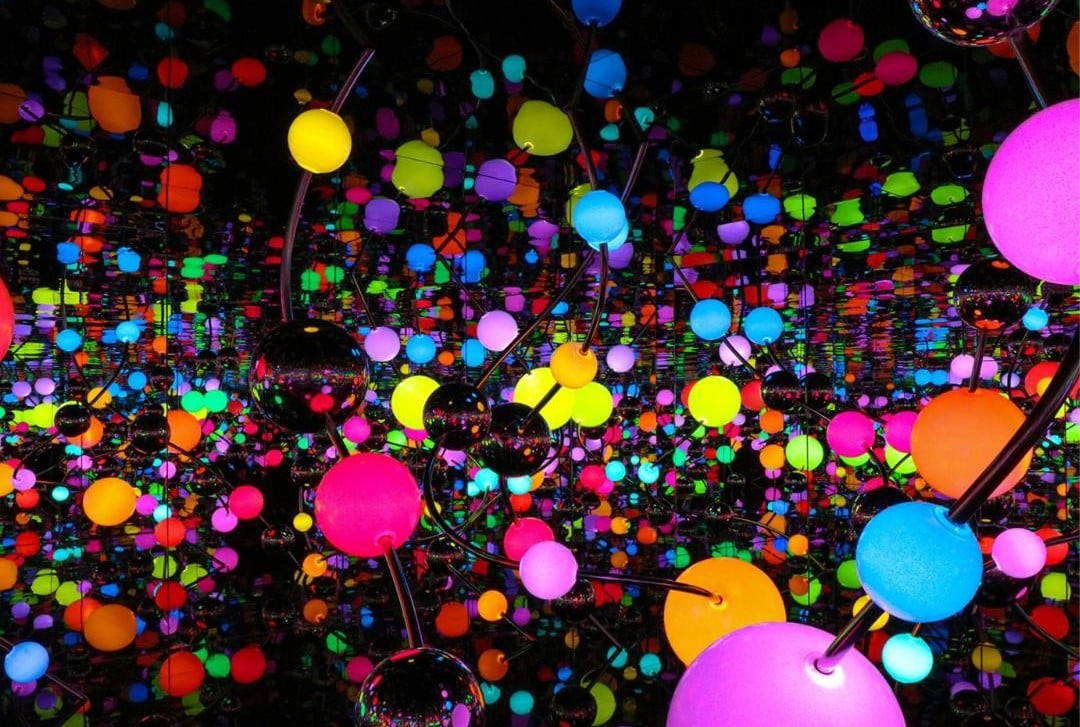 Colorful glowing spheres in neon shades of pink, yellow, blue, green, and orange are suspended among mirrored surfaces, creating a vibrant, endless reflection effect in a dark space.