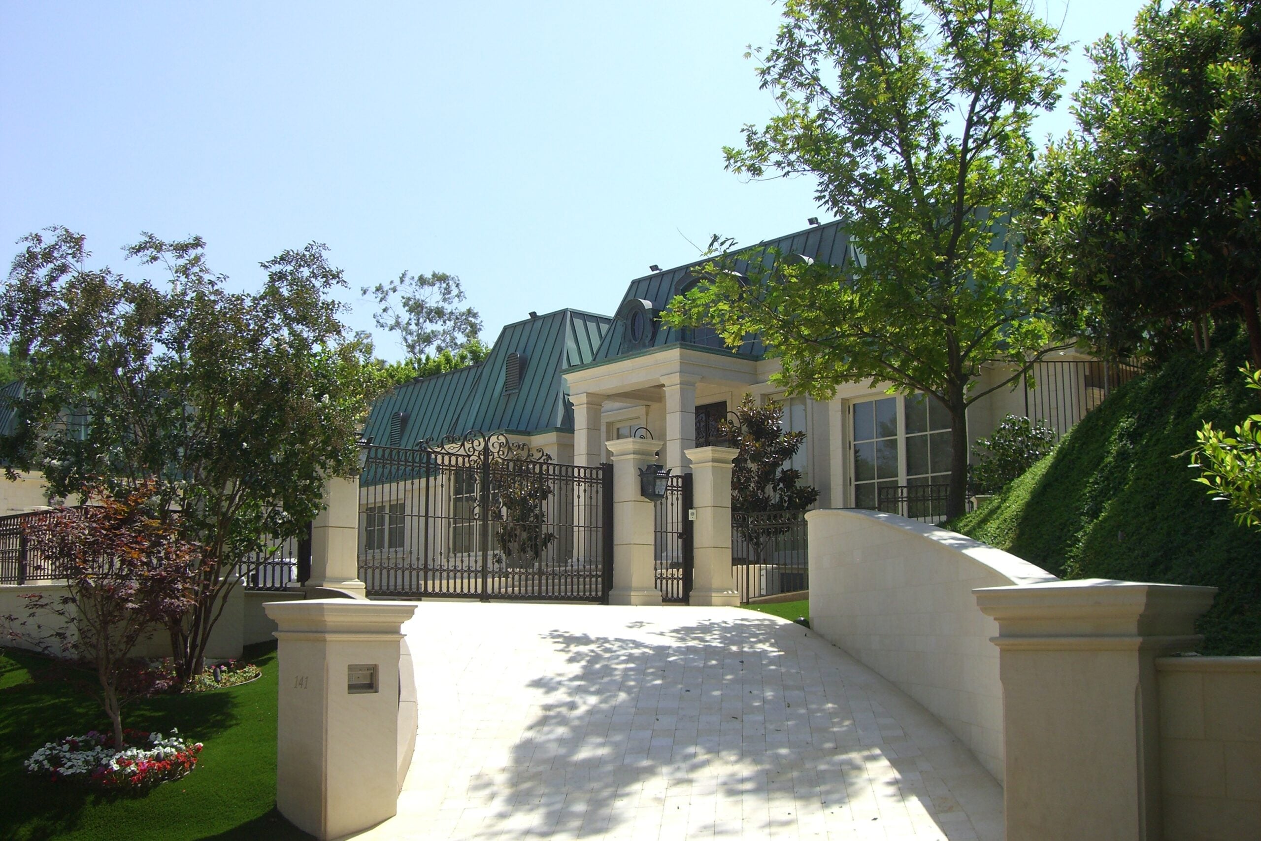 A large, elegant house with a green roof, beige exterior, tall columns, and black metal gate sits behind a steep, light-colored driveway, surrounded by trees and manicured landscaping on a sunny day.