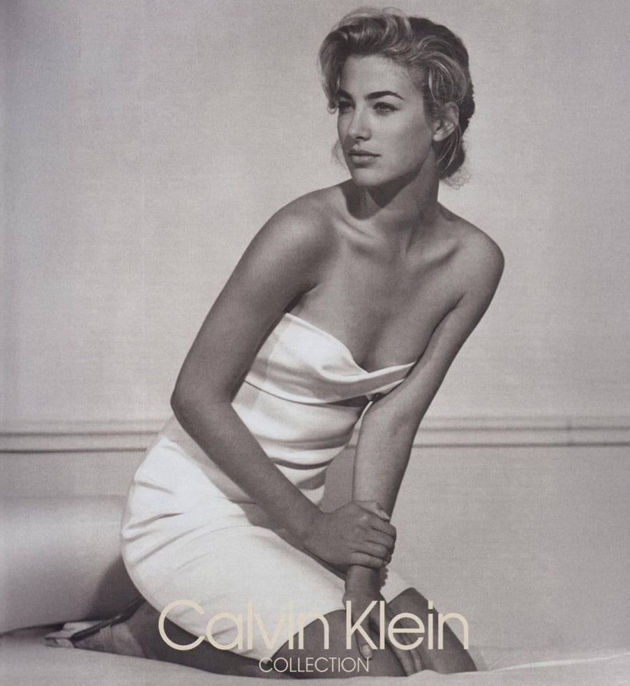A woman in a strapless white dress poses seated, looking to the side, against a minimal, light background. The image is in black and white with "Calvin Klein Collection" text at the bottom.