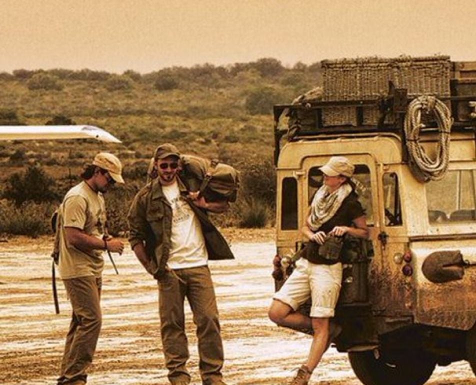 Three people in outdoor clothing stand by a dusty, rugged vehicle loaded with gear on a dirt road, with a plane and brushy landscape in the background. The scene has a warm, sepia-toned color.