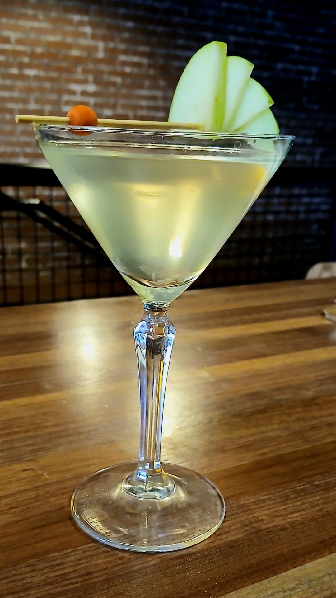 A clear martini in a stemmed glass garnished with three thin green apple slices and a cherry on a cocktail pick, placed on a wooden table with a blurred brick wall background.