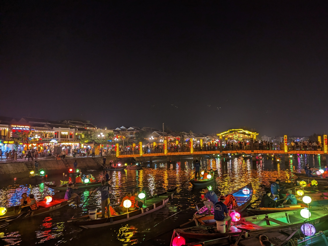 A lively night scene shows boats with glowing lanterns floating on a river, while crowds gather along a brightly lit waterfront filled with colorful lights and buildings.