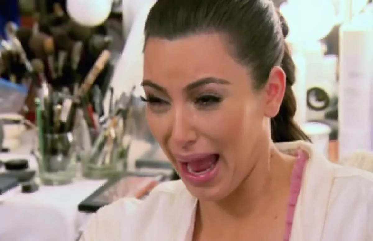 A woman with dark hair is crying with her mouth open and tears streaming down her face, sitting in front of a cluttered makeup table in a brightly lit room.