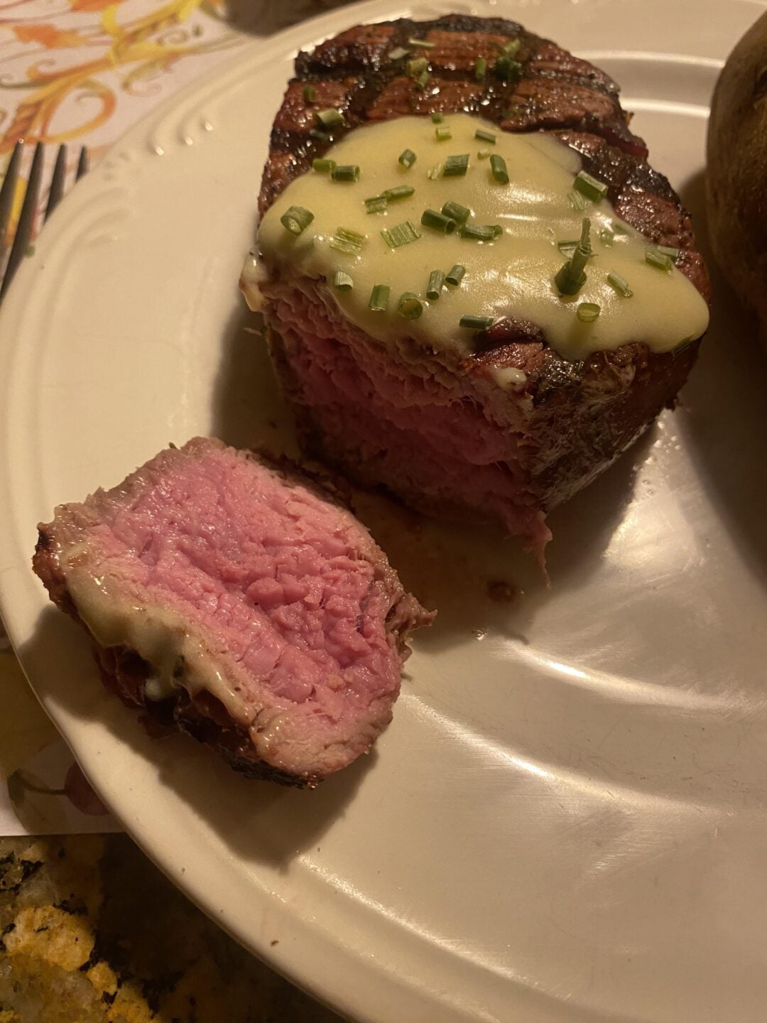 A thick, medium-rare steak with melted cheese and chopped chives on top sits on a plate. One slice has been cut and placed next to the steak, showing the pink interior. A baked potato is partially visible on the side.