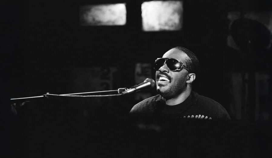 A man wearing dark sunglasses sings passionately into a microphone while playing the piano under dim stage lighting.