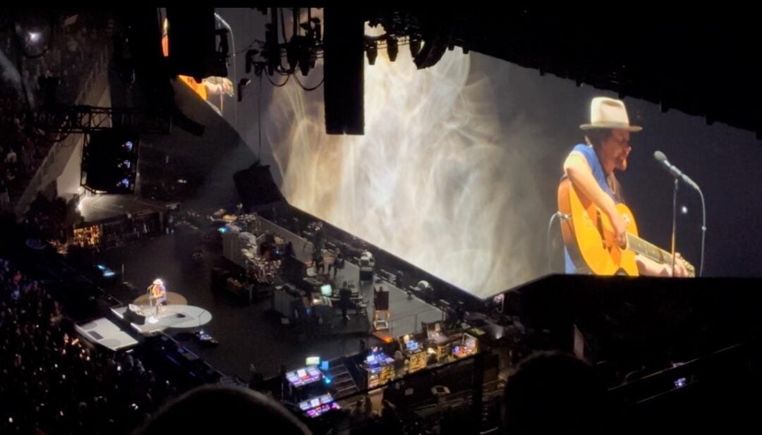 A musician wearing a hat plays an acoustic guitar and sings on stage at a large concert; his image is projected on a big screen behind him, with stage lights and audio equipment visible around the area.