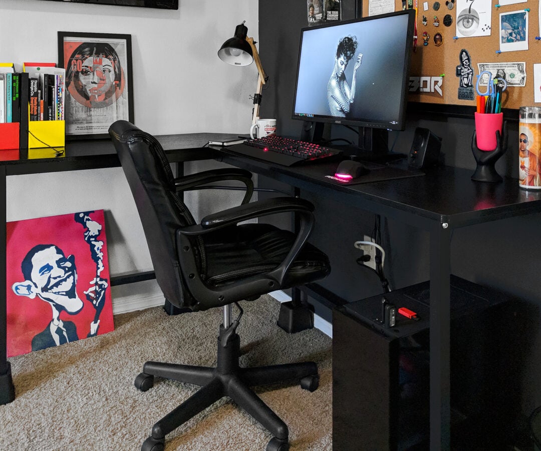 A modern home office features a black desk with a computer, keyboard, mouse, and lamp. The walls display colorful art, a corkboard with photos, and a mounted TV. A black chair sits on a carpeted floor.