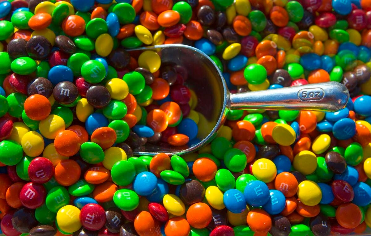 A metal scoop rests on a large pile of colorful candy-coated chocolate pieces, mostly green, blue, red, orange, yellow, and brown, with some displaying the letter "m.