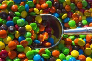 A metal scoop rests on a large pile of colorful candy-coated chocolate pieces, mostly green, blue, red, orange, yellow, and brown, with some displaying the letter "m.