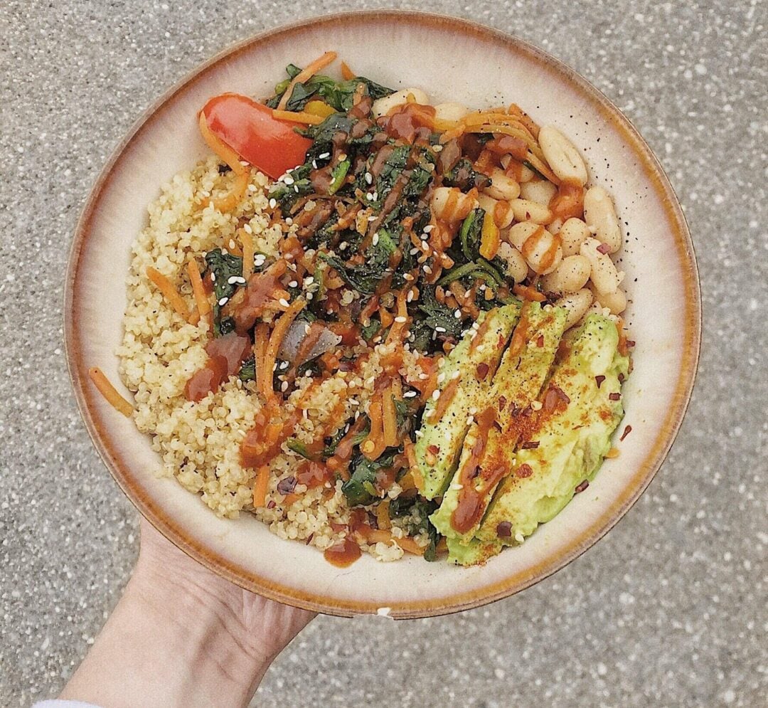 A hand holds a bowl filled with quinoa, avocado slices, white beans, sautéed greens, shredded carrots, a tomato wedge, and a drizzle of sauce, all topped with sesame seeds and red pepper flakes.
