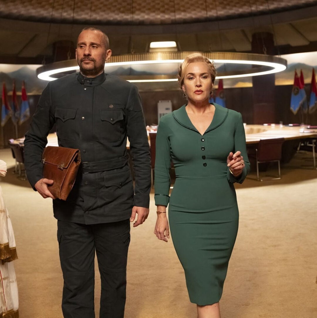 A man in a dark uniform holding a brown leather briefcase walks beside a woman in a fitted green dress inside a large, circular meeting room with flags and a glowing circular light overhead.
