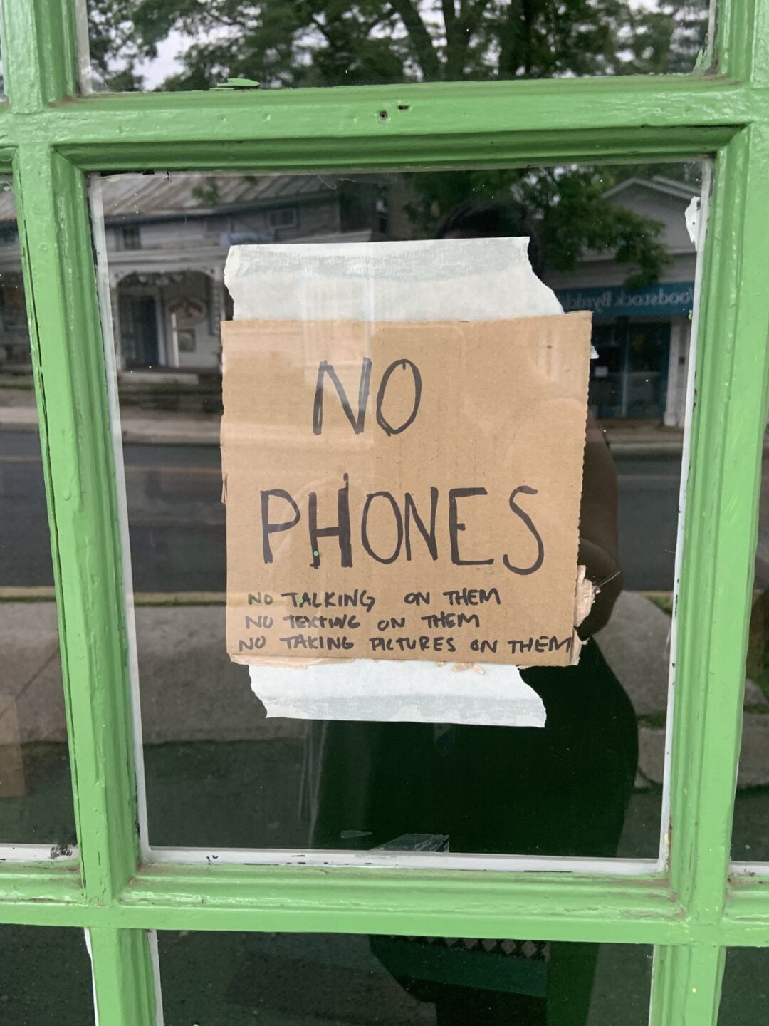 A handwritten cardboard sign taped to a green-framed glass door reads: "NO PHONES. No talking on them, no texting on them, no taking pictures on them." A street and buildings are visible outside.