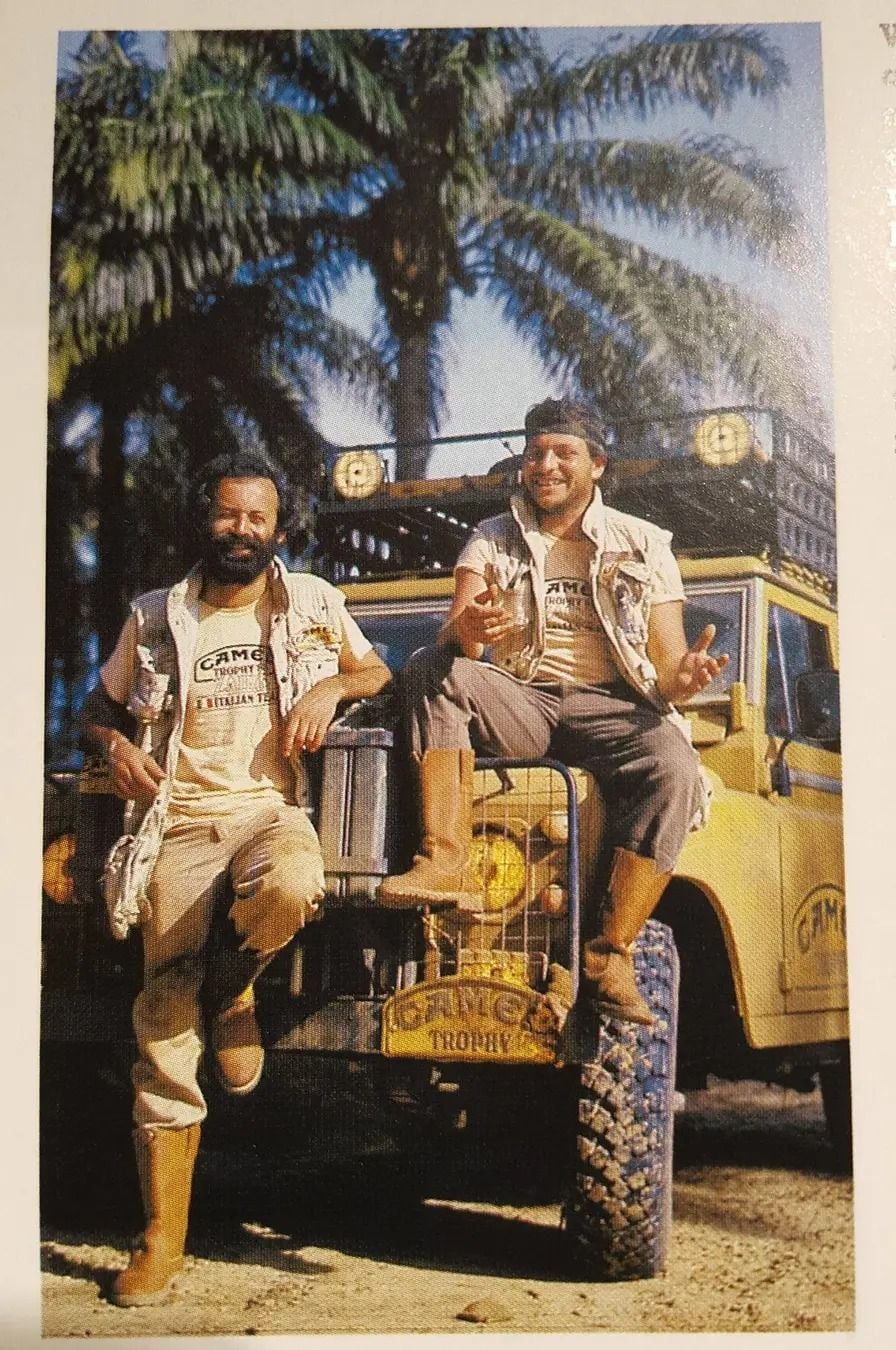 Two men in adventure gear smile and pose on the front of a yellow Land Rover with "Camel Trophy" branding, parked outdoors with tall palm trees in the background. One man sits on the hood while the other leans beside him.