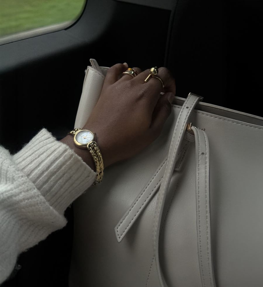 A person wearing gold rings and a watch rests their hand on a light beige handbag. They are dressed in a white sweater, and the scene appears to be inside a car.