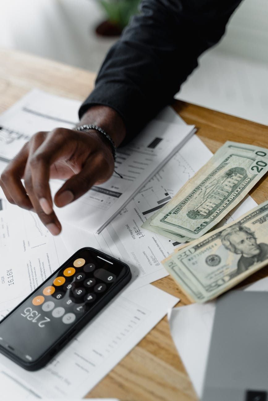 A person’s hand over scattered financial documents on a desk, next to a smartphone with a calculator app displaying 2365 and several US dollar bills.