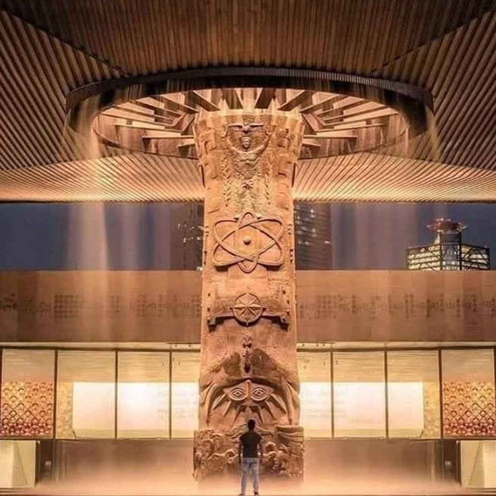 A person stands before a large, ornate stone pillar with intricate carvings under a circular canopy that releases a curtain of water, located at the National Museum of Anthropology in Mexico City.