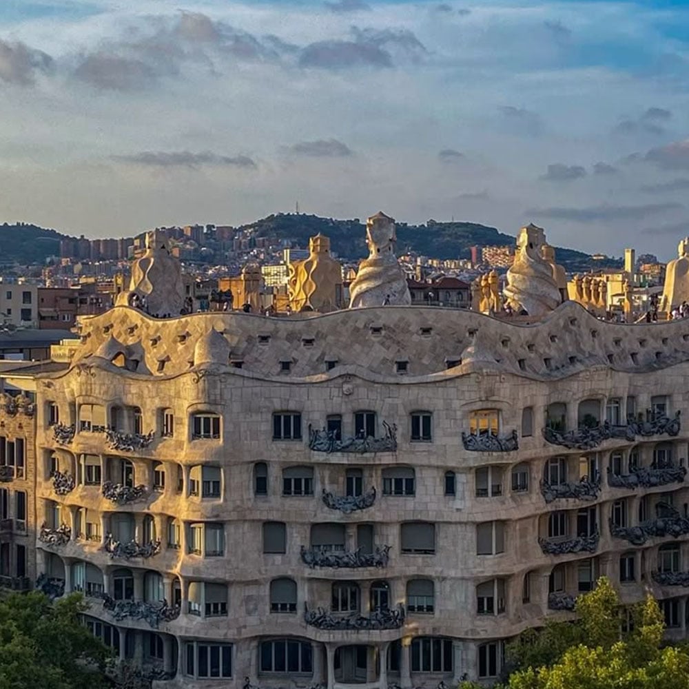 A wavy, stone building with sculpted chimneys and ornate balconies, known as Casa Milà or La Pedrera, stands in front of a cityscape and distant hills under a partly cloudy sky in Barcelona.